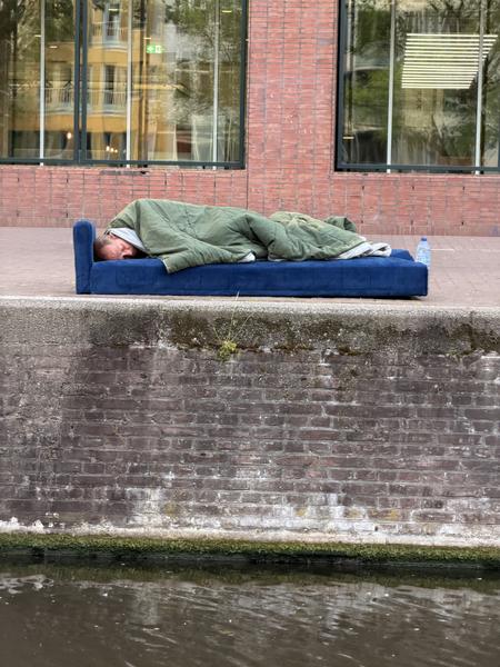 A person is seen sleeping on a blue mattress covered by a green blanket on the pavement next to a canal in Centrum, Amsterdam